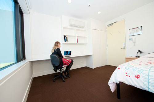 Female student on her laptop within her student accommodation, which consists of a single bed, desk and a chair. 