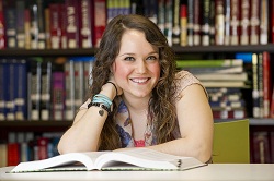  female smiling at camera in library with textbook in front of her, seated at desk