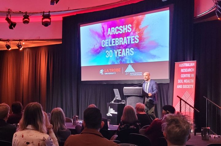 ARCSHS Acting Director Adam Bourne stands at a lectern on a stage in a function room, in front of a slide reading 'ARCSHS CELEBRATES 30 YEARS', next to a banner with 'Australian Research Centre in Sex, Health and Society' in front of an audience of people