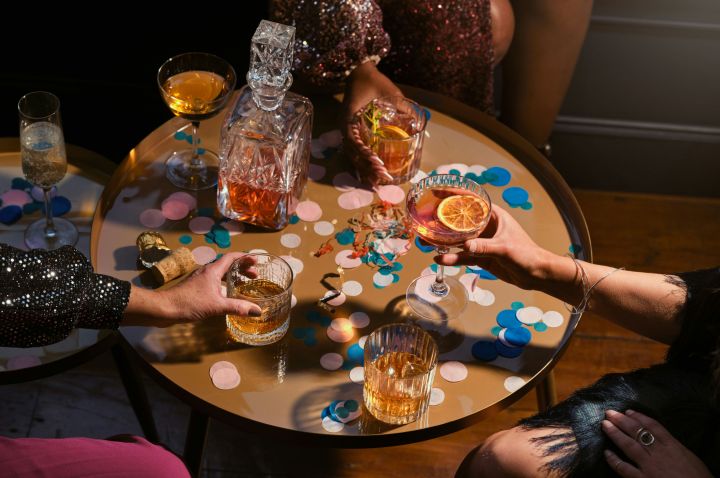 A table with a decanter of brown spirits, a sparkling wine cork, several glasses of sparkling wine and spirits, and large colourful confetti. Surrounding the table are three people's hands, each holding a drink, two wearing sparkly clothing.