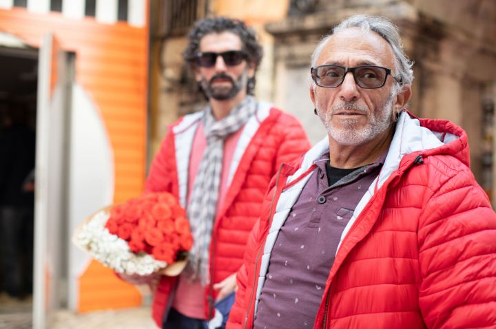 Two older men wearing matching red puffer jackets, one with a purple polo shirt and glasses, the other with a grey and white scarf and a large bunch of red and white flowers, from the ViiV HIV in View photo collection