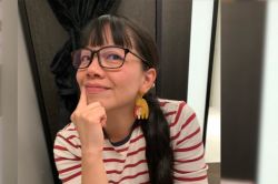 Dr Quah Ee Ling, a person with long black hair and a fringe, dark-framed glasses, a red and white striped top and an abstract shaped brightly-coloured large resin earring. She is smiling and holding a finger to the corner of her mouth.
