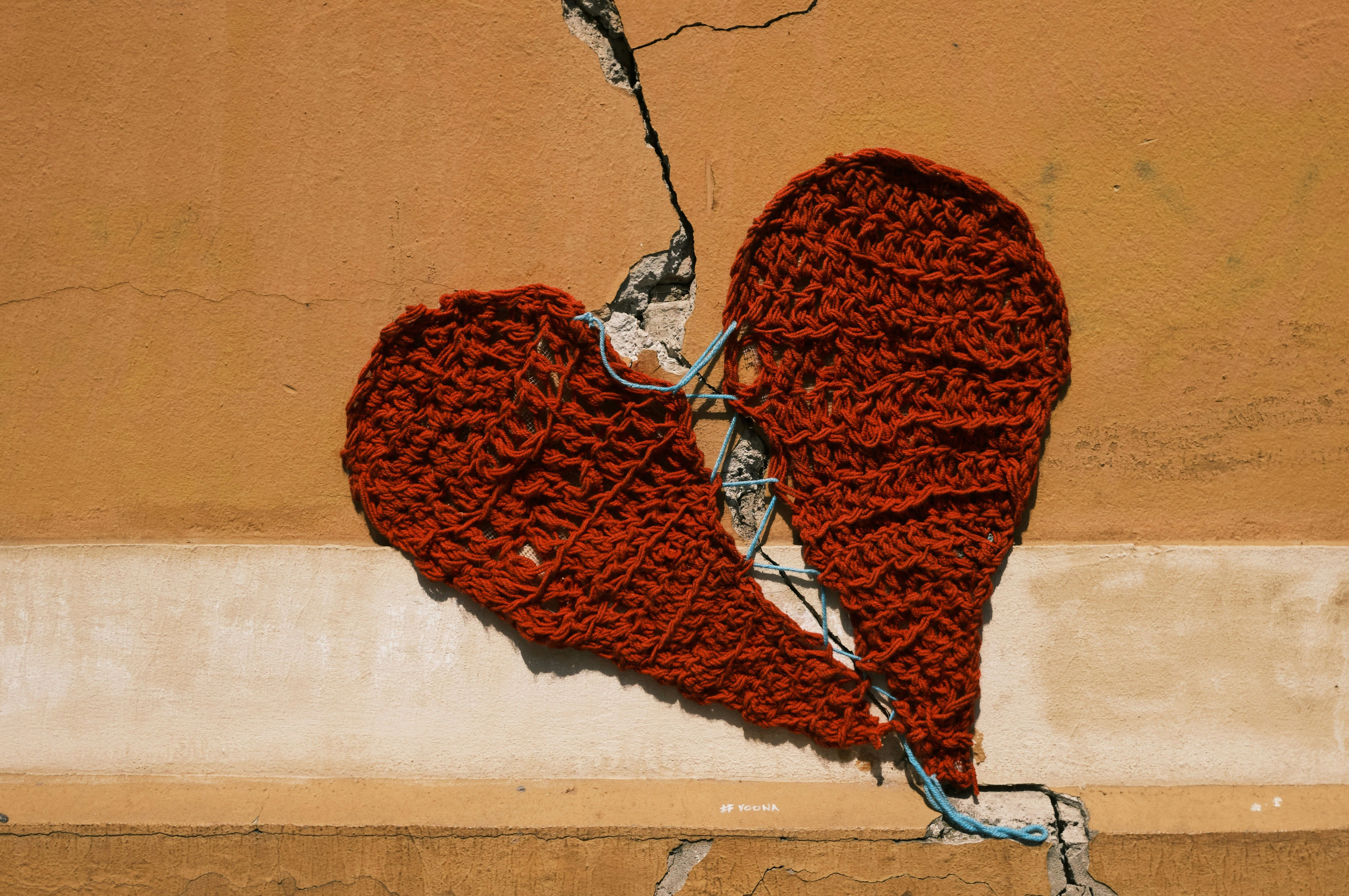 Two halves of a red crochet heart pasted either side of a diagonal crack in an orange wall. The two halves ha have been haphazardly sewn together with blue thread.