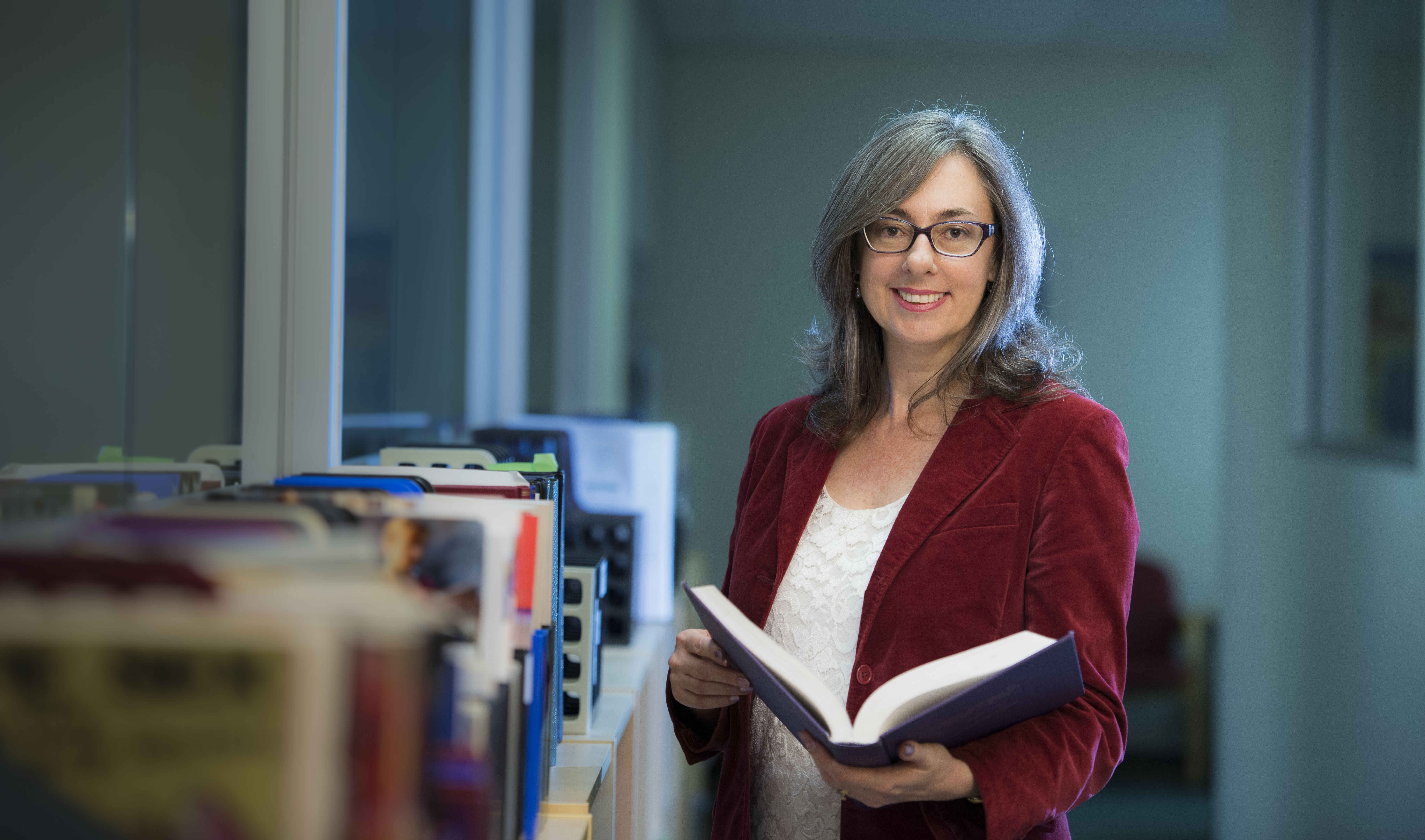 Photograph of Jayne Lucke, a person with long grey hair, glasses, a red velvet jacket and white top, holding a book and standing next to a bookshelf 