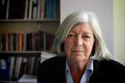 Photograph of Marian Pitts, a person with shoulder-length white hair, a blue shirt and black jacket, in front of a shelf of textbooks and folders