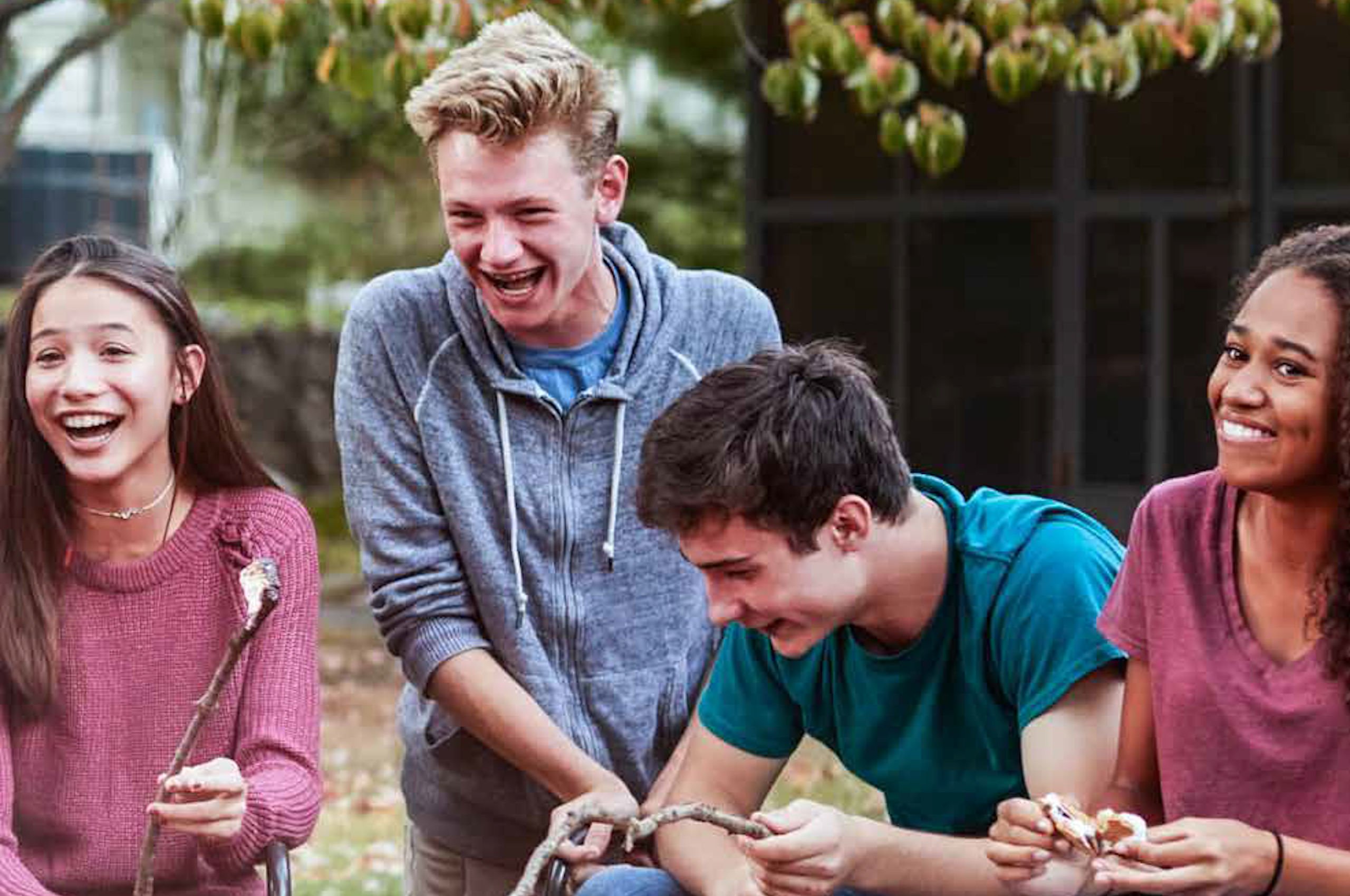 Four teenagers outdoors laughing and roasting marshmallows