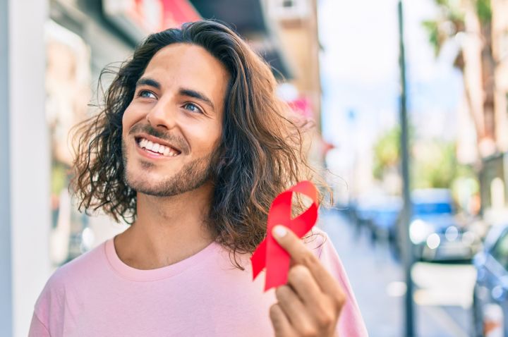 Man with long hair and pink T-shirt smiling while holding a folded red ribbon