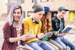 Four young people with colourful clothes and hair, sitting on a bench outside a school, looking at devices and books and smiling