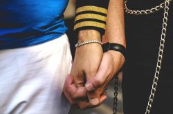 Close up of two people holding hands, one with white pants, a blue t-shirt and black watch, one with a black jacket with three gold pilot's stripes on the cuff, decorative belt chains and diamante bracelet