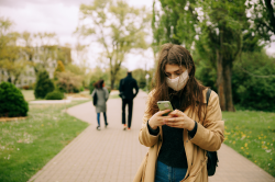 Woman in winter coat with long hair walks down pathway in garden while looking at phone