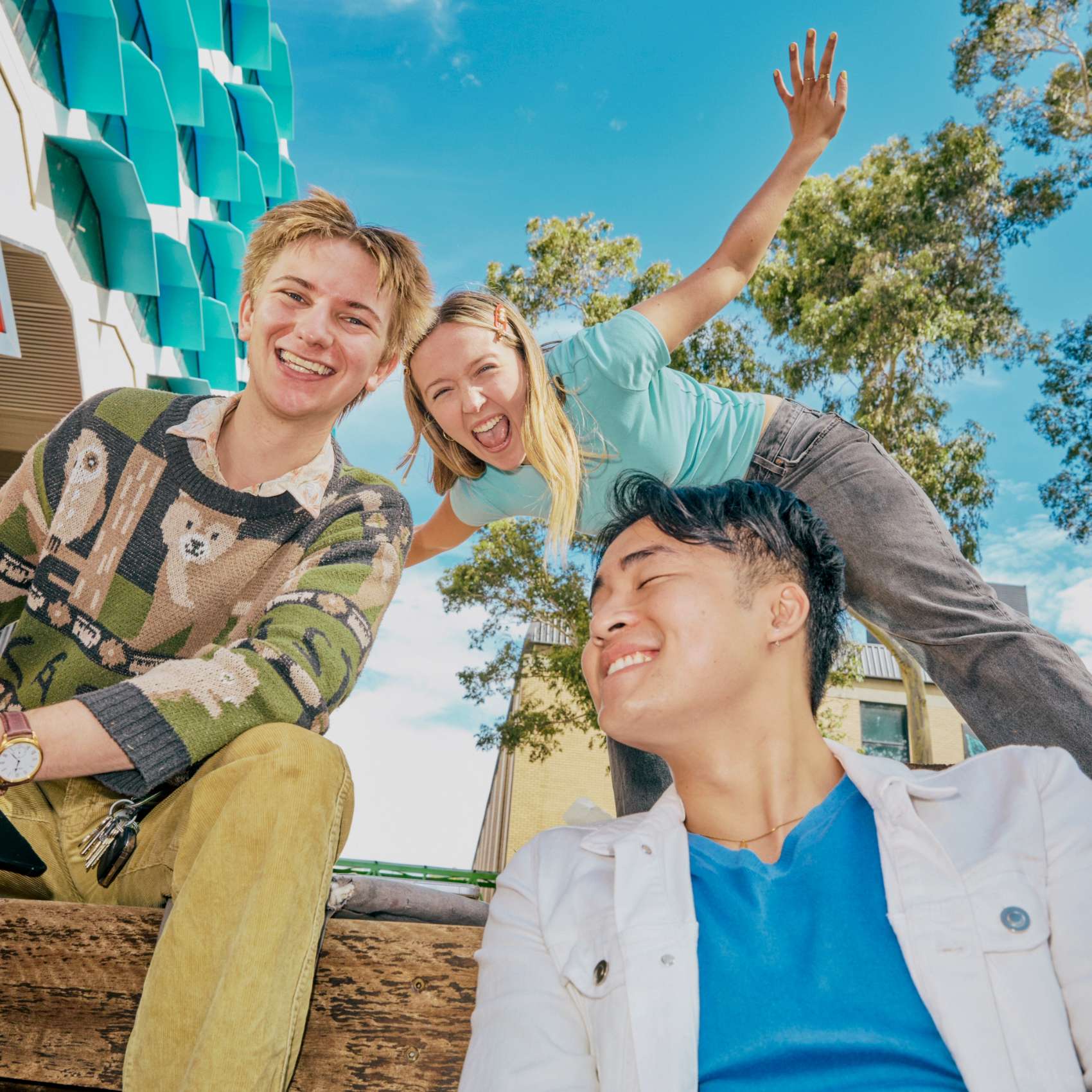 Three students laugh and pose together outdoors on a sunny university campus.