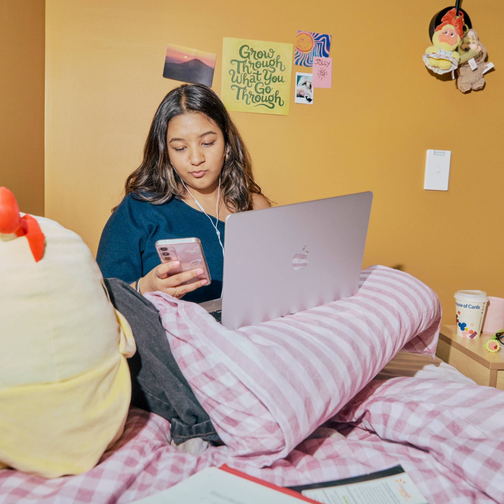 Student uses a laptop and phone while studying on a bed in a cozy dorm room.