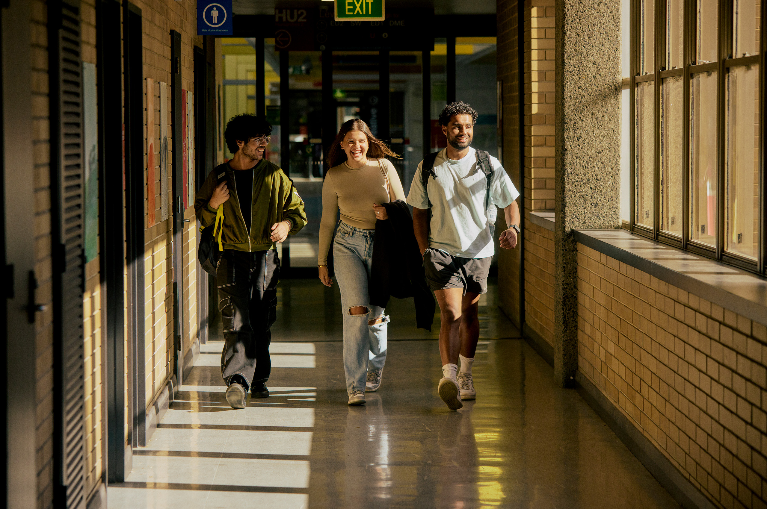 Three smiling students walking down a sunlit hallway.