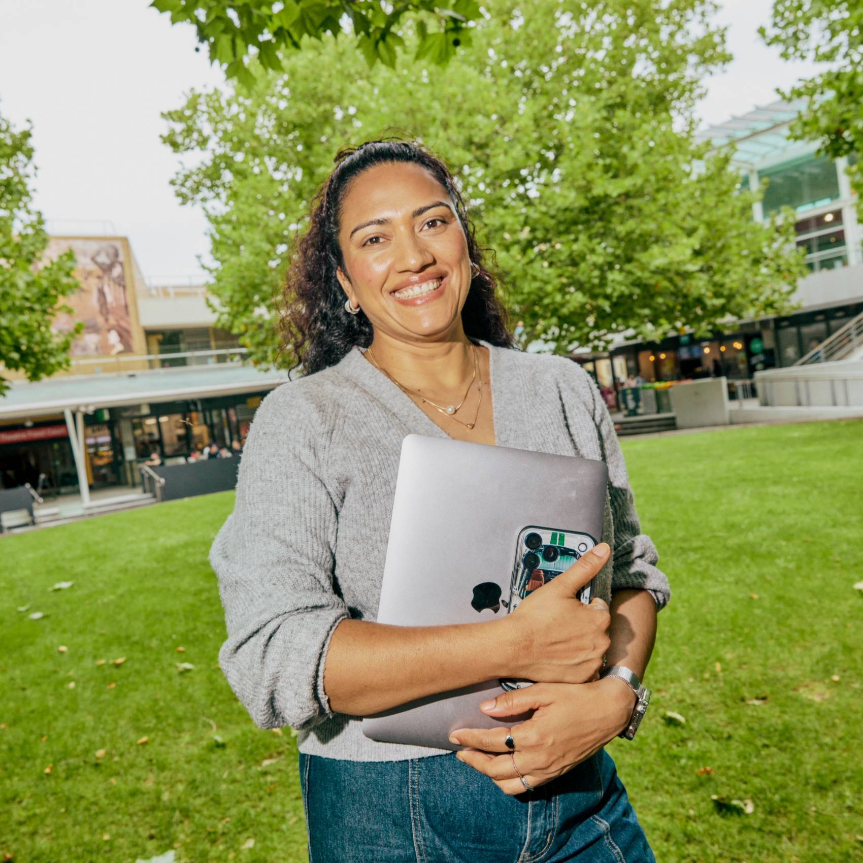 Woman smiles while holding a laptop and phone on a green university campus lawn.