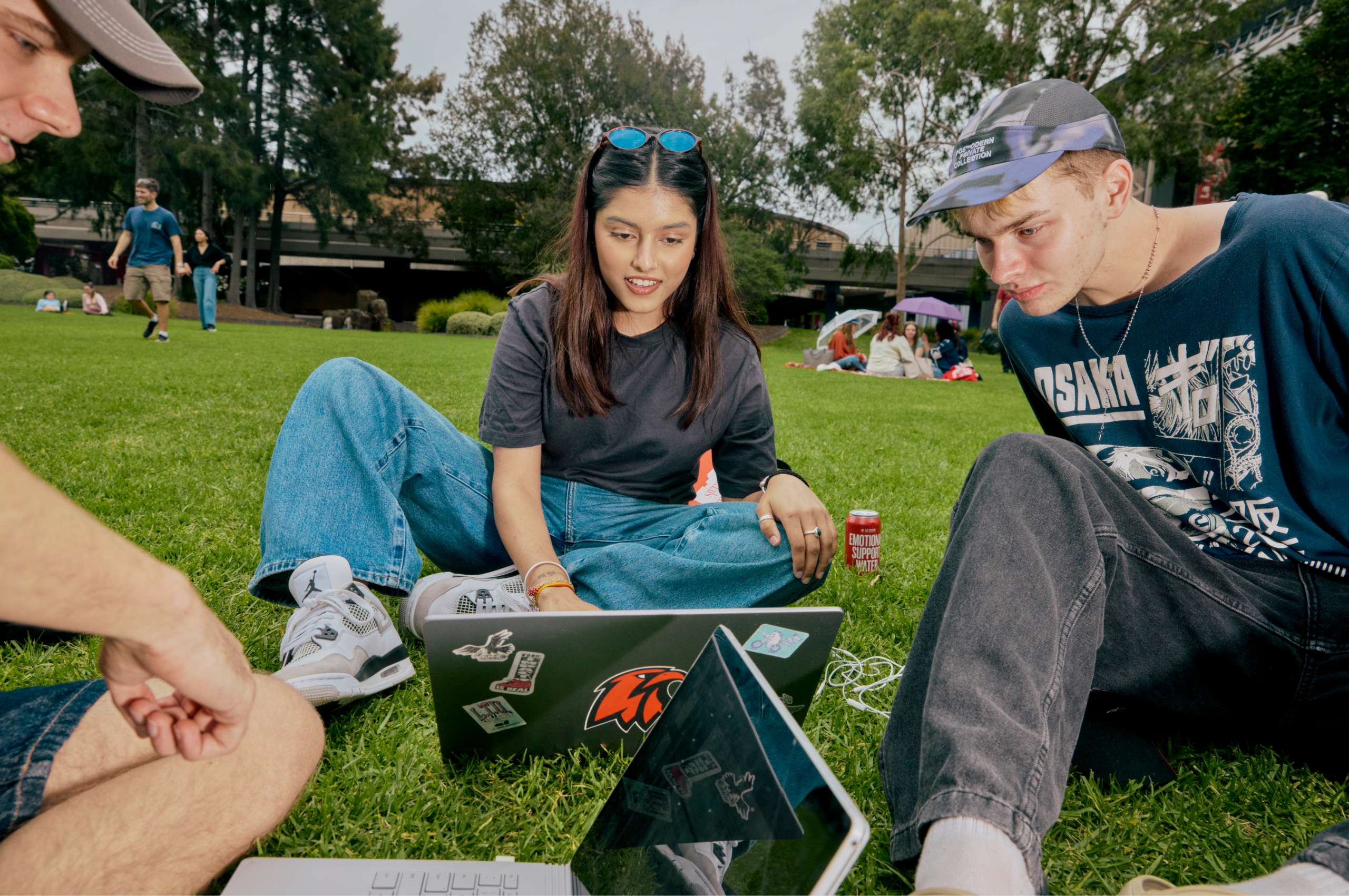 Young adults gather around laptops while sitting together on a grassy campus lawn.