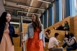 Students socializing and studying in a modern indoor common area with wooden tiered seating. 