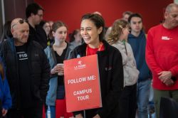 Female holding sign in crowded area. Sign reads: Follow me, campus tours.