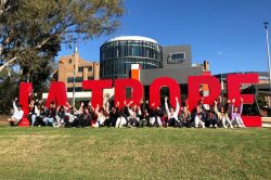 Students sitting on grass in front of large 'La Trobe' letters waving