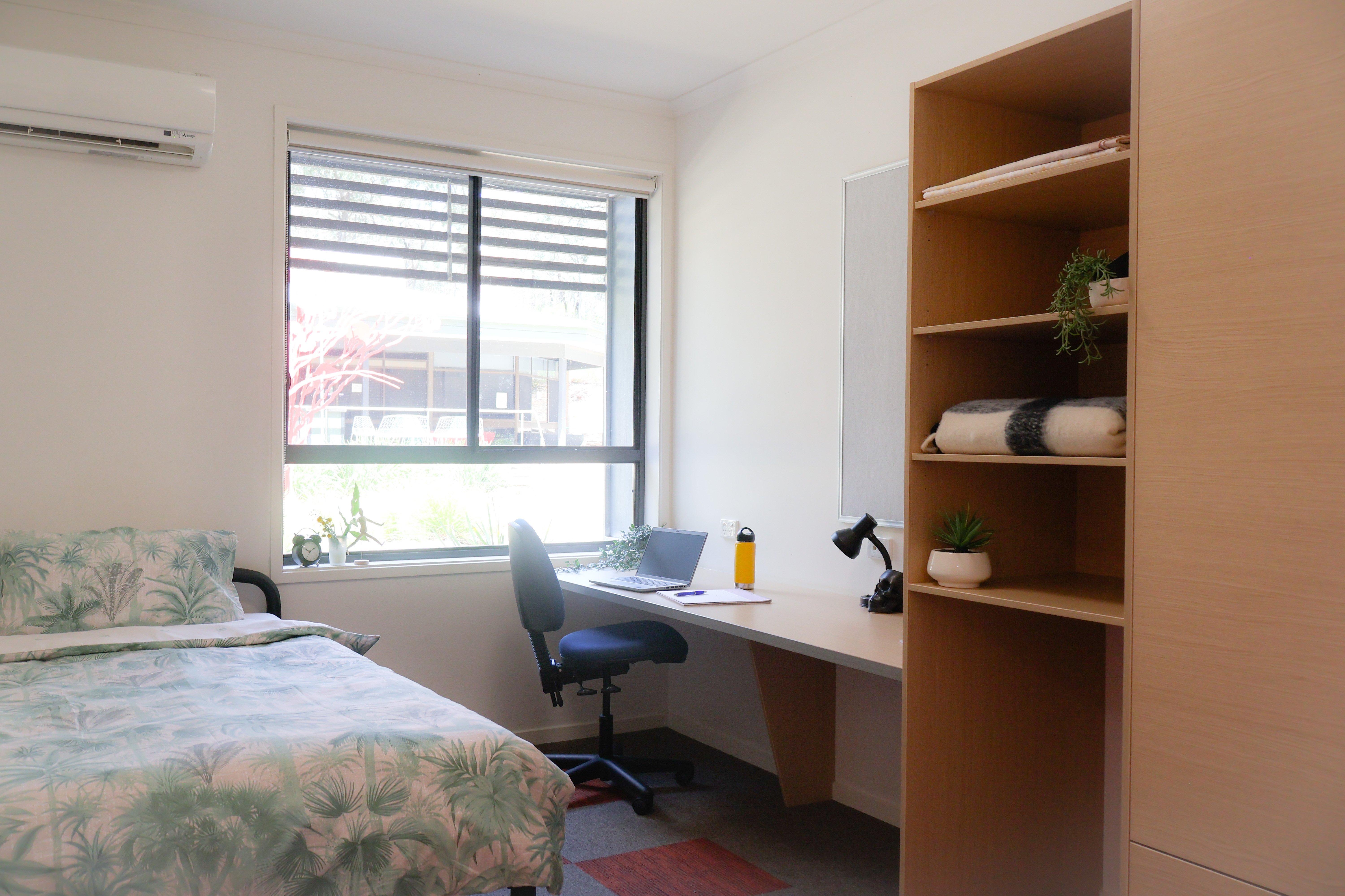 Single bed by bright window, built-in desk alongside open shelves and wardrobe