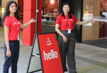 Two women in red text standing beside a sign that says "hello". The women are pointing towards a building, which is the Library at the Bundoora Campus.