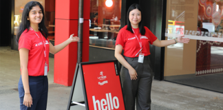 Two women in red text standing beside a sign that says "hello". The women are pointing towards a building, which is the Library at the Bundoora Campus.