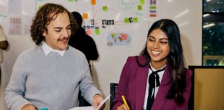 Two students sitting at table smiling. One is holding a pen and one is holding a piece of paper