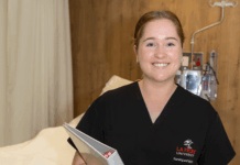 A student in a nursing uniform holding a file next to a hospital bed