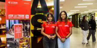 Two student advisors standing in front of the Ask La Trobe signs in the library at Bundoora
