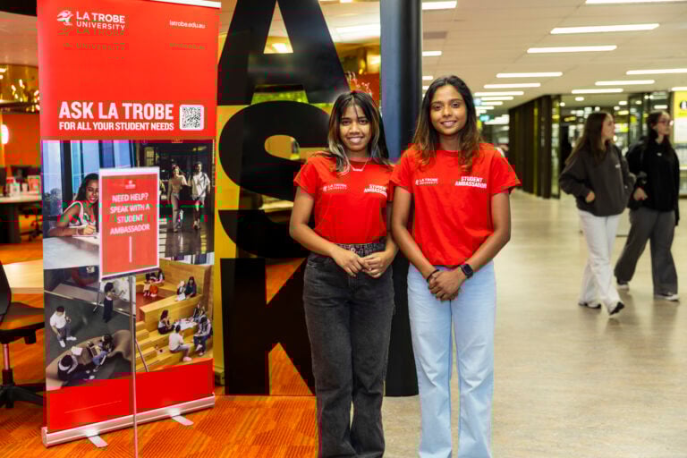 Two student advisors standing in front of the Ask La Trobe signs in the library at Bundoora