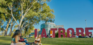 Students sitting on the grass by a large La Trobe letter sign