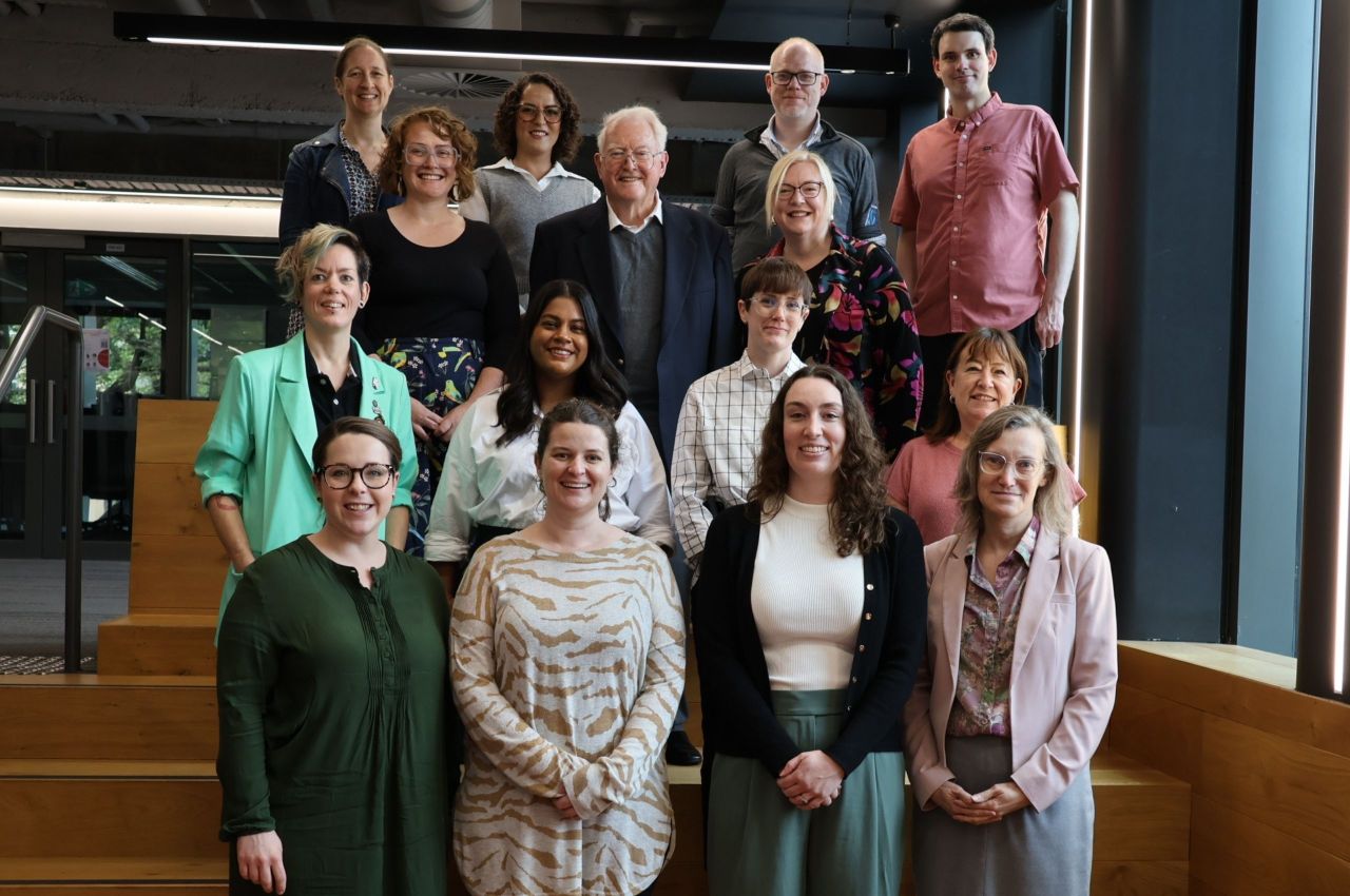 A group of people stands together in front of a large window, smiling and engaging with one another