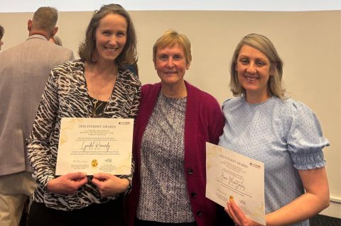 Three women proudly display certificates in front of a large screen, celebrating their achievements together