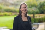 A woman in a suit stands confidently in front of a lush green park, showcasing a professional yet relaxed demeanor
