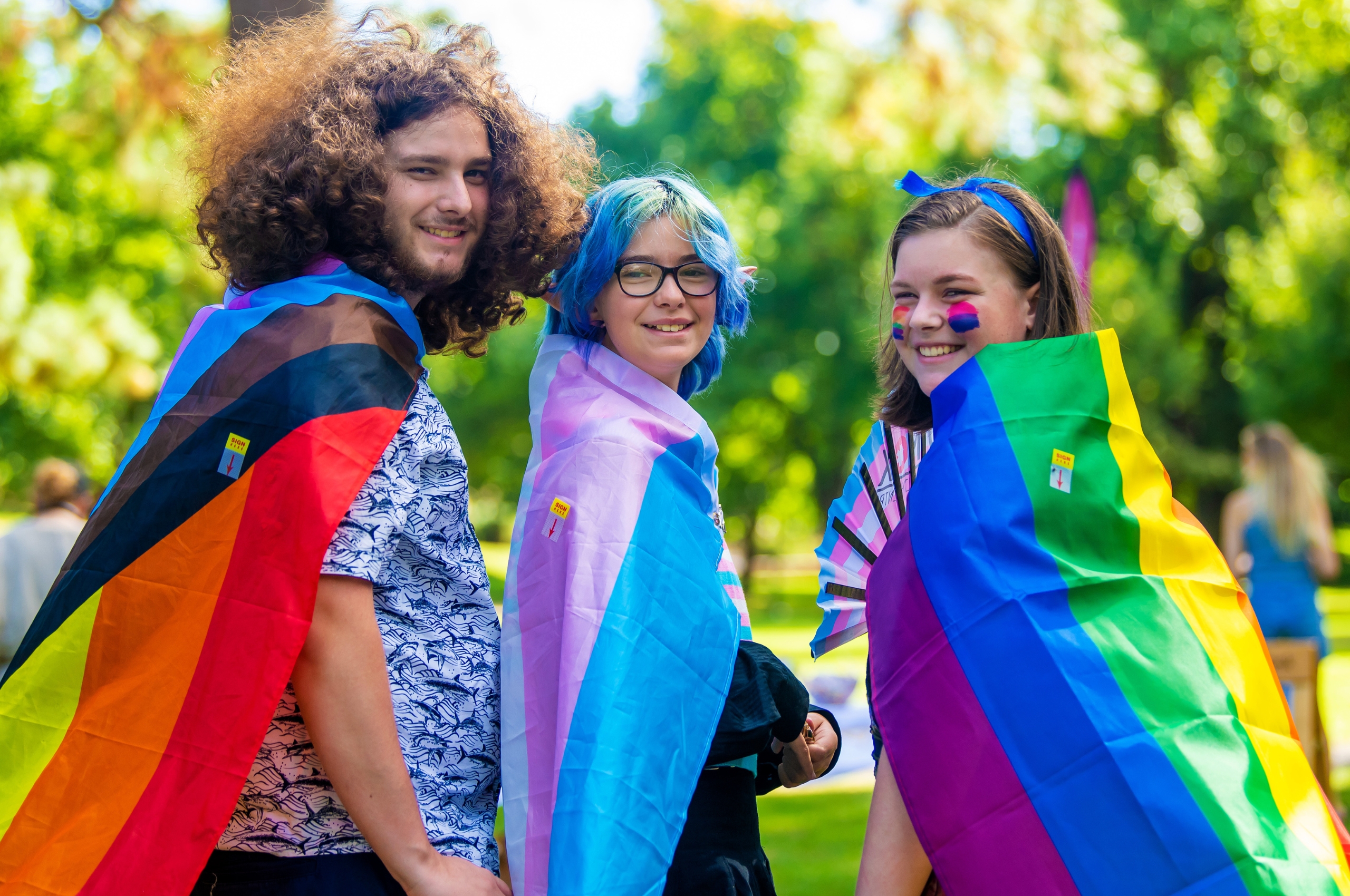 Three students at an LGBTIQA+ celebration, wearing pride flags around them
