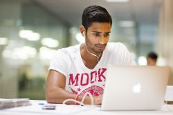 male student sitting in library looking at laptop with headphones in at desk