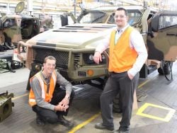 2 males in reflective vests smiling at camera standing in front of military camouflage vehicle 