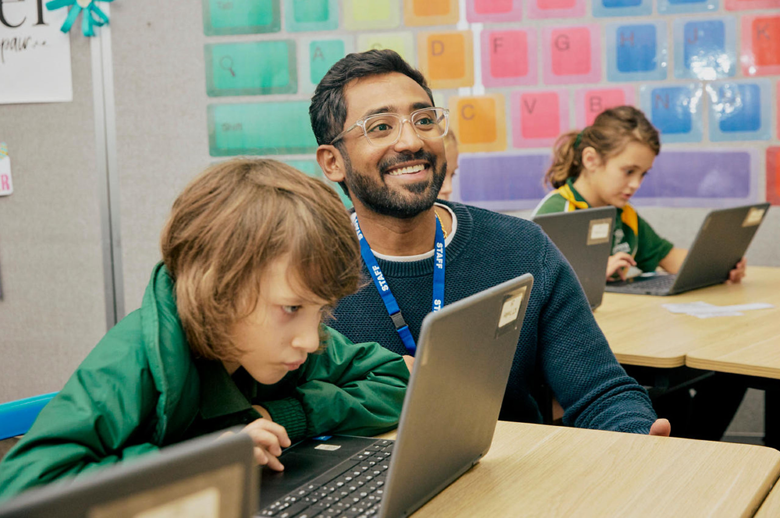 A male teacher sitting next to a primary school boy in a classroom.